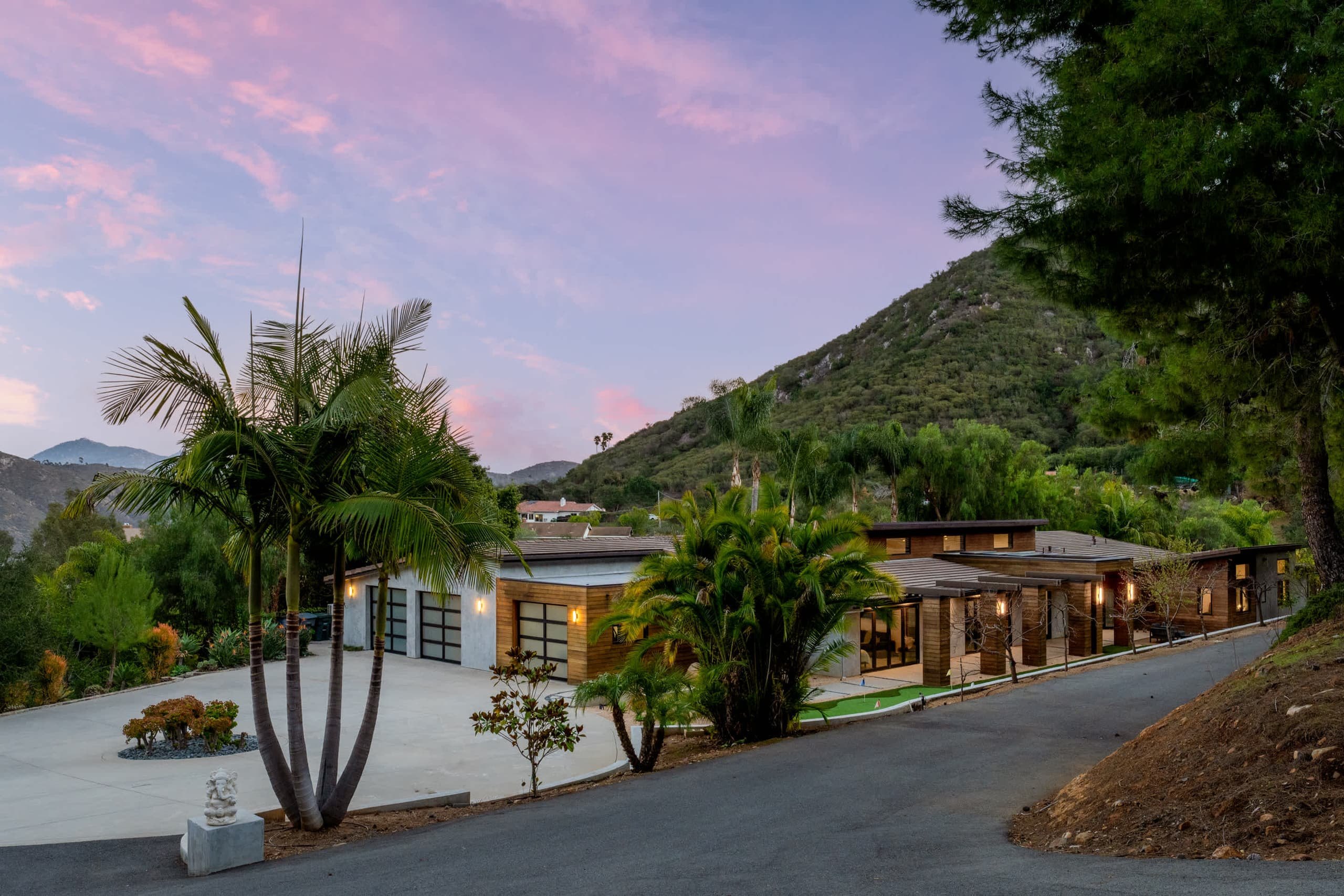 Palm-lined drive leading to the lit house at dusk, hills behind