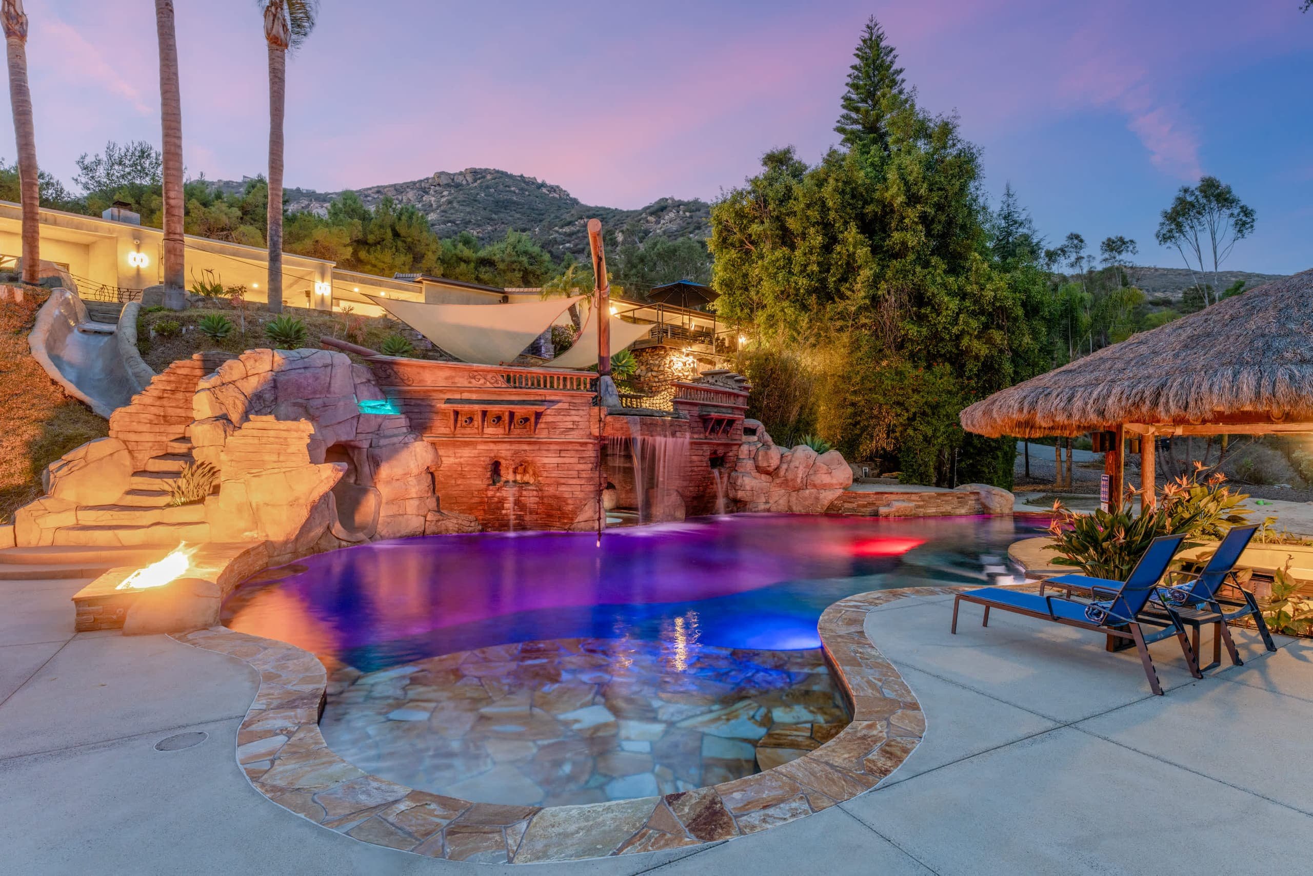 Pool at dusk with rock grotto, slide, fire pit, palms and blue loungers